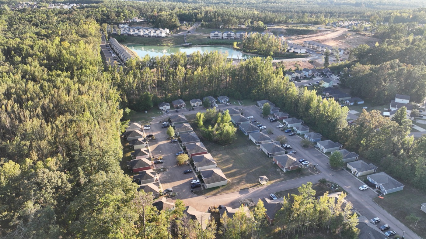 High drone view of the Acadia Loop homes surrounded by pine woods, with a pond and more housing beyond, in Oxford, MS