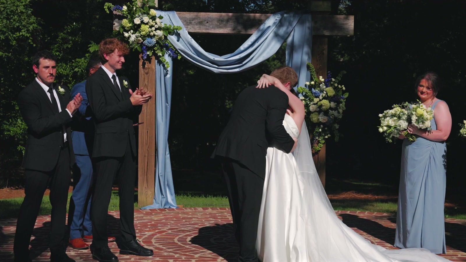 Bride and groom embracing under the same draped arch as the groomsmen clap and a bridesmaid holds her bouquet