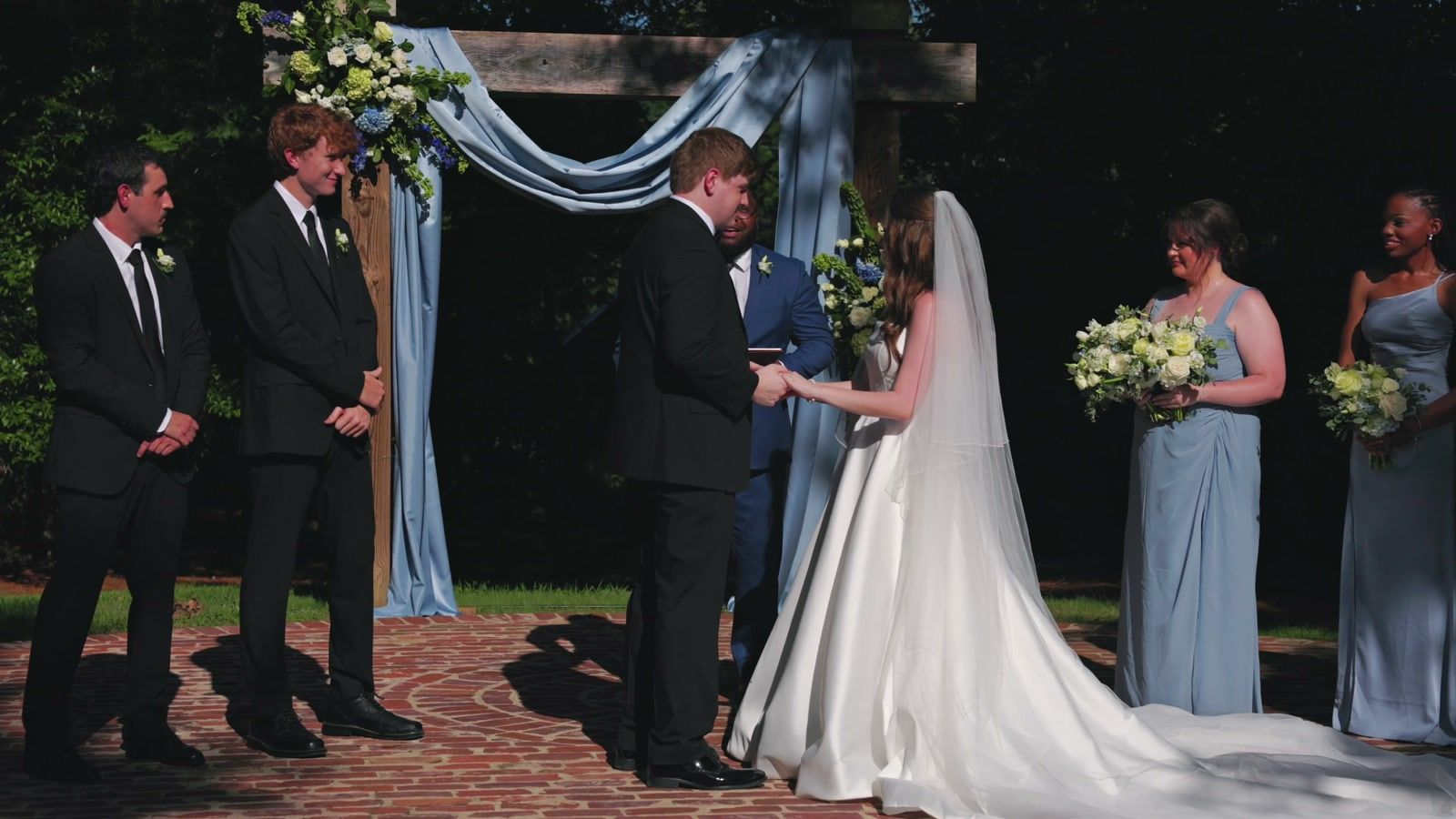 Bride and groom holding hands under a wooden arch draped in blue fabric, groomsmen in black suits and bridesmaids in light blue on either side, on a brick patio