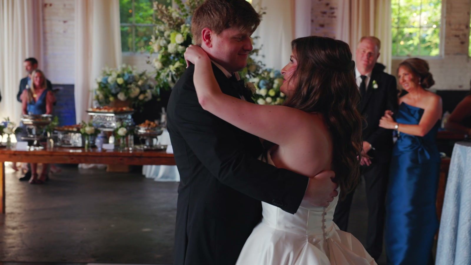 Bride and groom dancing close in a reception hall with whitewashed brick walls as family watches beside a food table