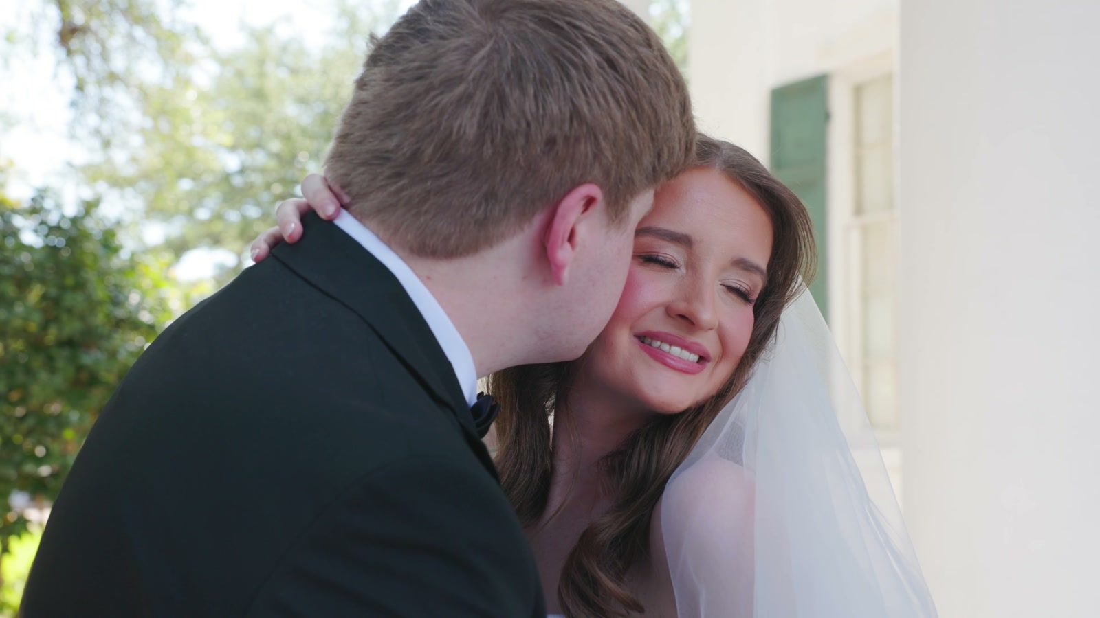 Close shot of the groom kissing the bride's cheek as she smiles with her eyes closed, outdoors in daylight