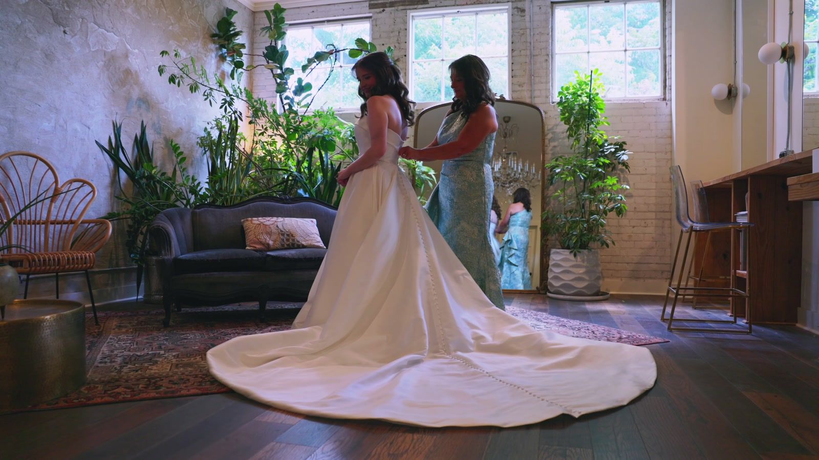 Bride standing in her gown with a long train in a sunlit room full of plants while a woman in a blue dress helps with the back of the dress